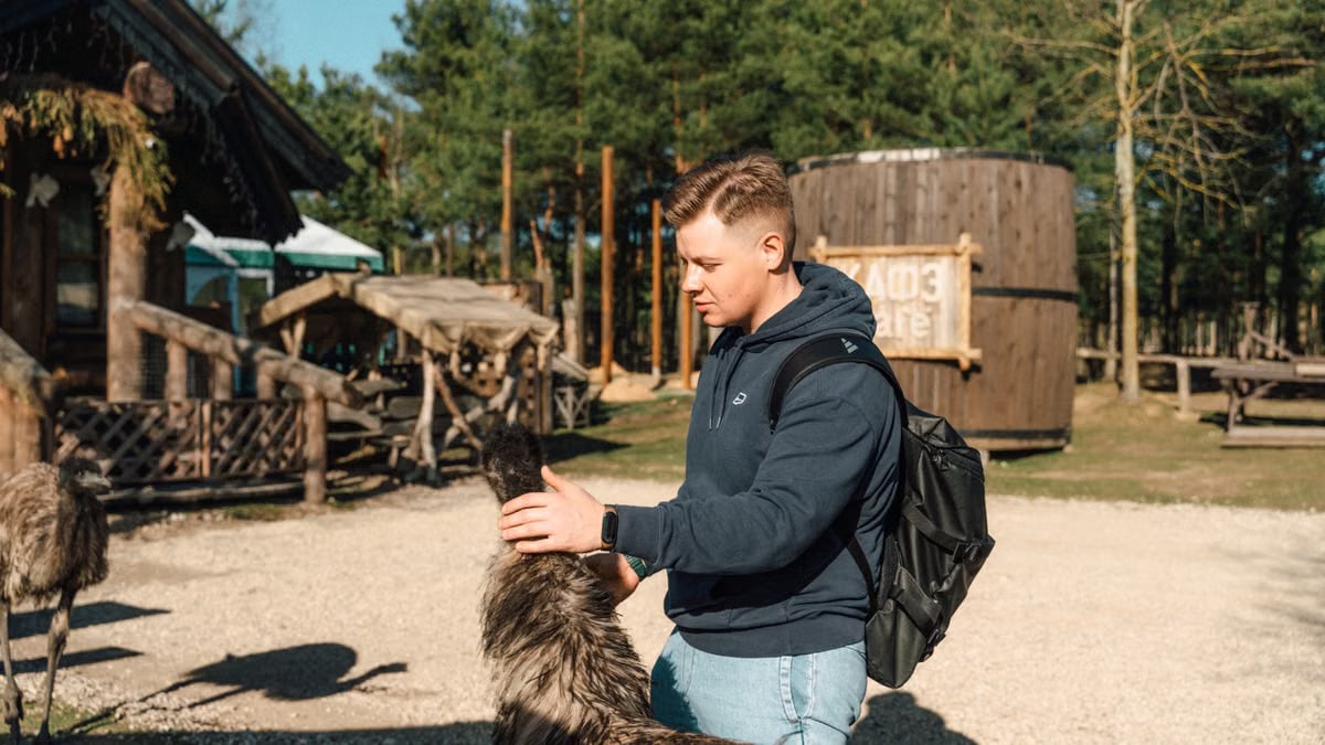 A young man with a backpack pets an emu on a sunny day in a rustic outdoor area.